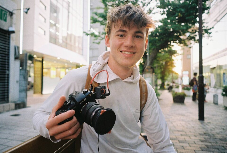 Grant Hall sitting on a bench outside, holding a GH5, wearing a white hoodie, and carrying a backpack